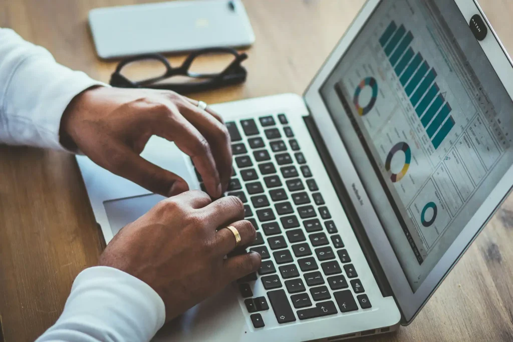 In the picture, a close-up of a person's hands working on a laptop. A phone and a pair of glasses rest on the desk.