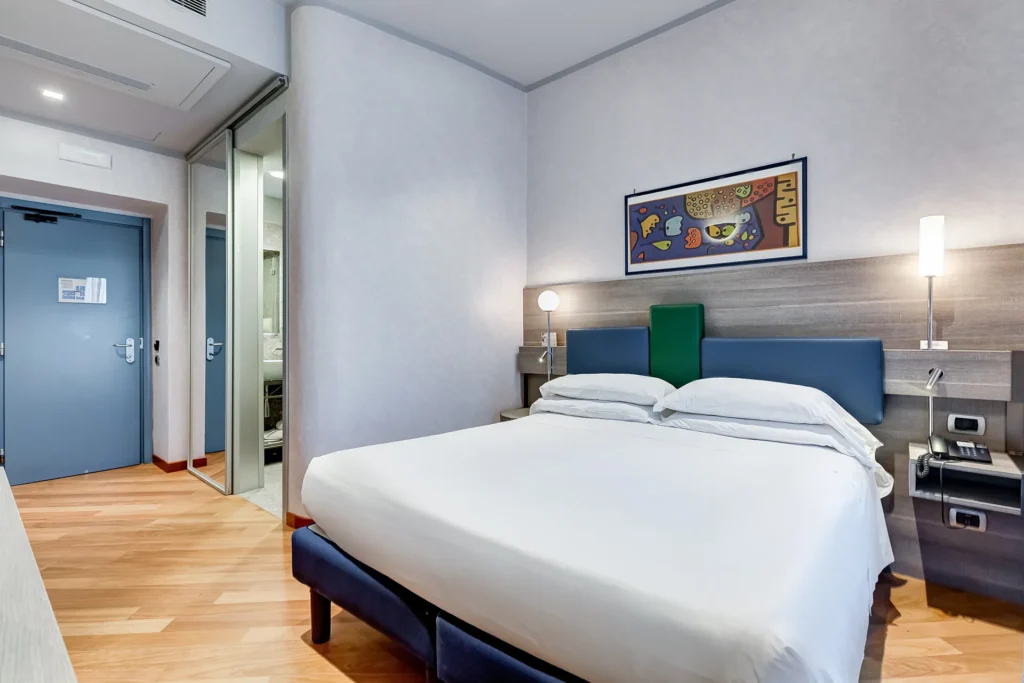 Nell'immagine, la camera Matrimoniale Superior dell'Hotel Plaza: si vede il letto matrimoniale e, sulla sinistra, si intravede il bagno attraverso la porta aperta. Di fianco, c'è uno specchio a figura intera appeso alla parete e la porta d'ingresso della stanza.