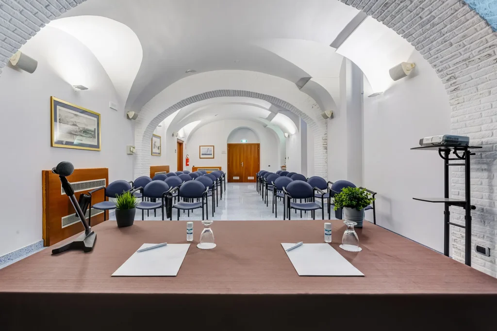 In the picture, the Principe Meeting Room of the Hotel Plaza, from the speakers' perspective. In the foreground, we can see the table with paper and pens, two glasses, and a microphone. In front, several rows of chairs.